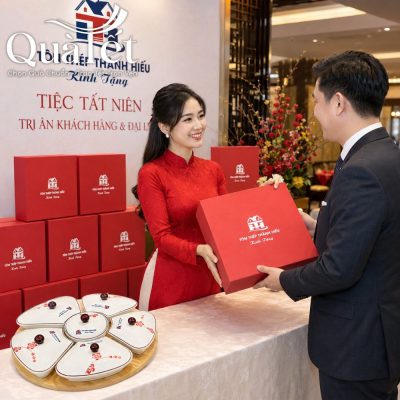Lễ tân trong trang phục áo dài đỏ trao tặng hộp quà Tết in logo cho khách hàng tại sự kiện tất niên