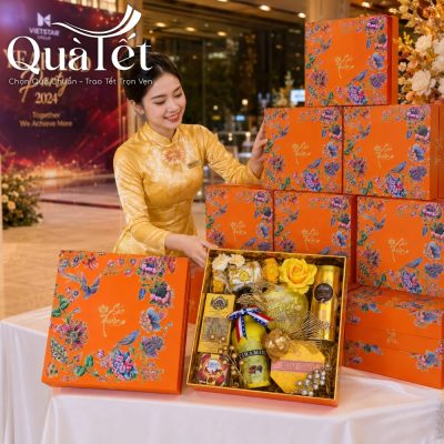 Nữ nhân viên mặc áo dài lụa vàng đang xếp các hộp quà Tết Sắc Xuân màu cam thành chồng ngay ngắn tại sự kiện Year End Party 2024 của doanh nghiệp