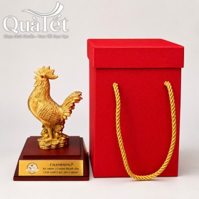 Cận cảnh tượng gà vinh danh mạ vàng QT-TG01 đặt cạnh hộp đựng lót lụa màu đỏ có quai xách dây thừng xoắn